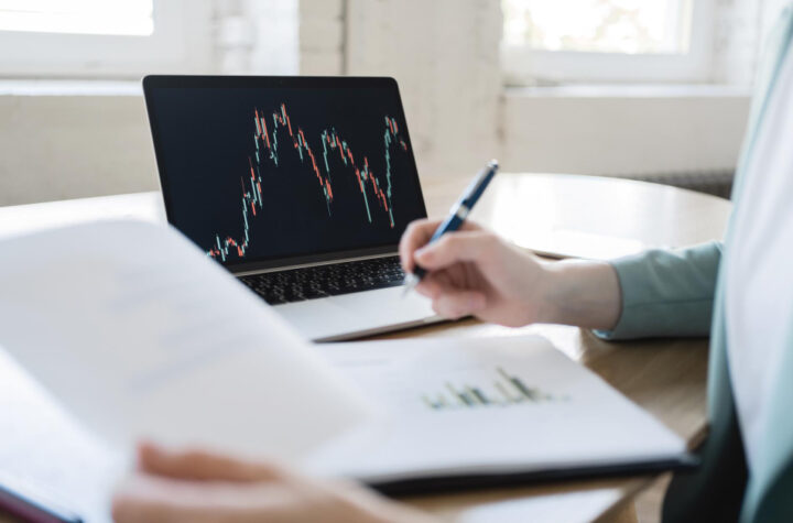 A person is studying financial charts on a laptop screen while holding a pen and reviewing a document, suggesting an analysis of market trends or investment research.